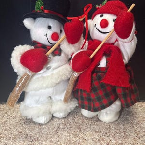 Könnte beinhalten: Zwei Schneemannfiguren. Einer trägt einen schwarzen Zylinder und der andere eine rote Strickmütze. Beide haben rote Fäustlinge, Schals und Nasen. Sie halten Holzbesen und sind in weiß-roten Karomustern gekleidet, ideal für die Weihnachtsdekoration.