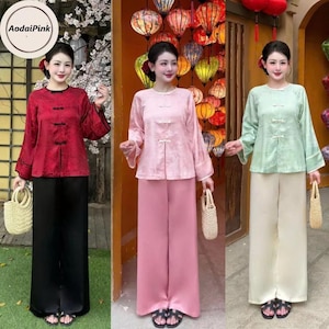 May include: Three women wearing traditional Asian-style outfits. The outfits include long-sleeved tops with frog closures and wide-leg pants. Colors include red and black, pink, and light green and cream. Each woman holds a woven bag.