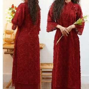 May include: Two women wearing matching red dresses. The dresses have a textured, floral-like pattern and long sleeves. The dresses are floor-length and paired with matching pants. One woman holds flowers.