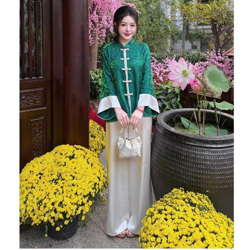 Op de afbeelding: Een groen en wit traditioneel Aziatisch ge&iuml;nspireerd outfit. De top is groen met een bloemenpatroon en witte accenten, gecombineerd met een cr&egrave;mekleurige broek. Een kleine, witte handtas wordt vooraan vastgehouden. De achtergrond bevat gele bloemen en een grote pot met een lotusbloem.