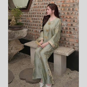 May include: A light green silk pajama set with gold embroidery. The set includes a long-sleeved top with a V-neck and wide-leg pants. The outfit is worn with white shoes and is set against a brick wall.
