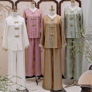 May include: Four sets of traditional-style clothing on mannequins. The outfits include long-sleeved tops and wide-leg pants in white, lavender, gold, and sage green. Each top features decorative gold clasps and trim.