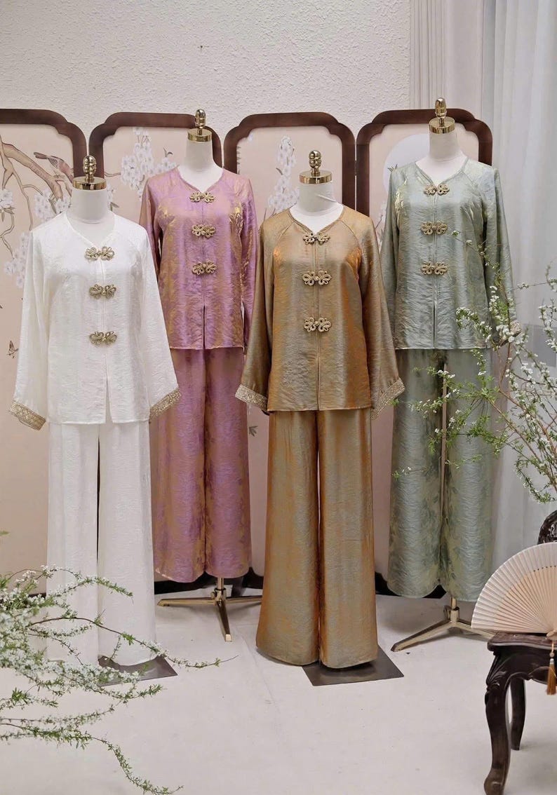 May include: Four sets of traditional-style clothing on mannequins. The outfits include long-sleeved tops and wide-leg pants in white, lavender, gold, and sage green. Each top features decorative gold clasps and trim.