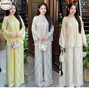 Op de afbeelding: Drie vrouwen in bijpassende sets traditionele kleding. De sets bestaan uit tops met lange mouwen en knopen en broeken met wijde pijpen. De outfits zijn in tinten geel, grijs en crème, allemaal met een bloemenpatroon. Elke vrouw draagt een kleine, zilveren handtas.