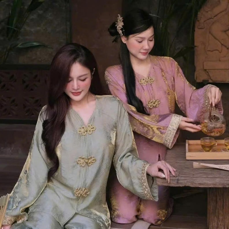 May include: Two women in traditional Asian attire. One wears a sage green top with gold embellishments, the other a pink top with gold trim. They are engaged in a tea ceremony, with a teapot and cups on a wooden table.