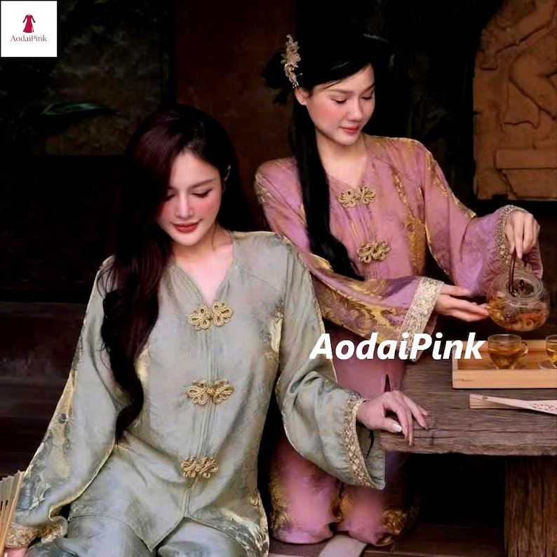 May include: Two women wearing traditional Asian-style silk outfits. One outfit is a light green color, and the other is a pinkish-purple color. Both outfits have gold-colored decorative clasps. The outfits have long sleeves and are made of a shiny material. The text "AodaiPink" is visible.