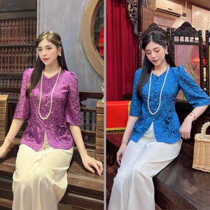 May include: Two blouses, one purple and one blue, with floral patterns and three-quarter sleeves. Each blouse has a V-neck, a row of buttons, and a pocket detail. The blouses are paired with white trousers and pearl necklaces.