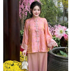 Op de afbeelding: Een roze traditionele outfit met een mandarijnkraag en kikkerknopen. De top met lange mouwen en de bijpassende broek zijn gemaakt van een textuurstof. De outfit wordt gedragen met een kleine zilveren tas.