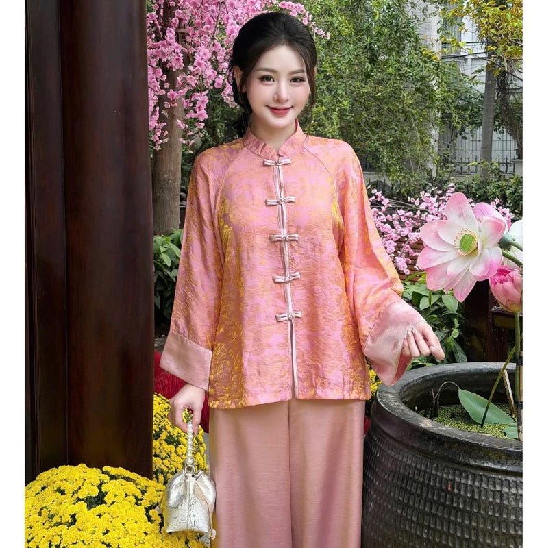 Op de afbeelding: Een roze traditionele outfit met een mandarijnkraag en kikkerknopen. De top met lange mouwen en de bijpassende broek zijn gemaakt van een textuurstof. De outfit wordt gedragen met een kleine zilveren tas.