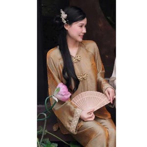 May include: A woman wearing a gold-colored silk outfit with floral button closures and embroidered cuffs. She holds a wooden fan and has a flower in her hair. A pink lotus flower is in the foreground.
