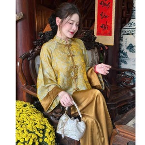 Op de afbeelding: Een vrouw in een goudkleurige traditionele outfit met een bloemenpatroon. De outfit bestaat uit een top met lange mouwen met kikkerknopen en een bijpassende broek. Ze houdt een witte tas met trekkoord en een parelhandvat vast.