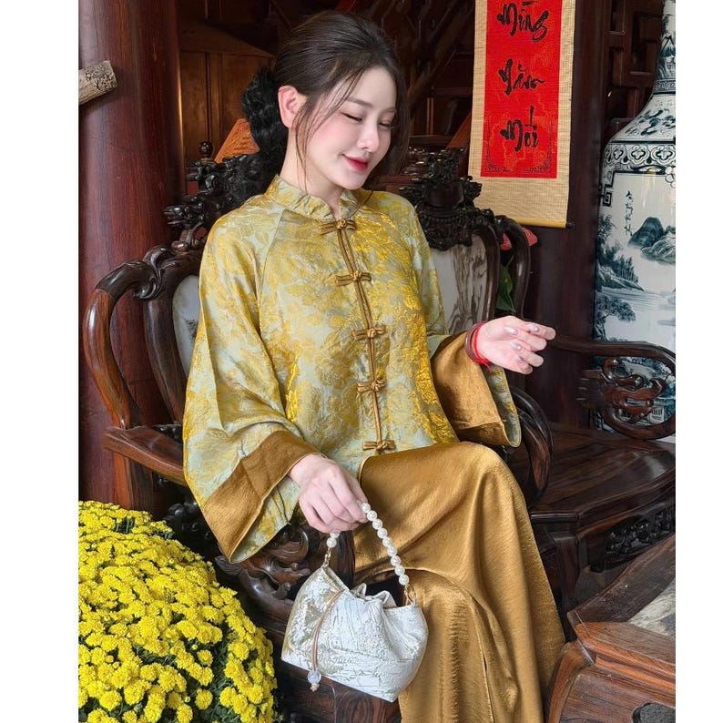 Op de afbeelding: Een vrouw in een goudkleurige traditionele outfit met een bloemenpatroon. De outfit bestaat uit een top met lange mouwen met kikkerknopen en een bijpassende broek. Ze houdt een witte tas met trekkoord en een parelhandvat vast.