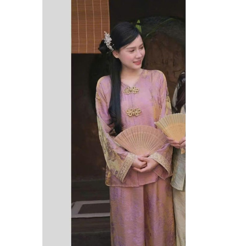 May include: A woman wearing a traditional pink outfit with gold accents, including a long-sleeved top and matching pants. The top features decorative gold clasps and the sleeves have ornate gold trim. She holds a tan fan.