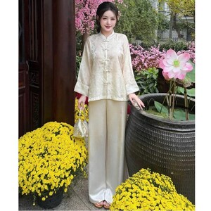 Op de afbeelding: Een cr&egrave;mekleurige tweedelige outfit met een top met lange mouwen, een mandarijnkraag en kikkerknopen, gecombineerd met een broek met wijde pijpen. De outfit heeft subtiele geborduurde details. Het model houdt een kleine, zilveren handtas vast. Op de achtergrond gele bloemen en een grote lotusbloem.