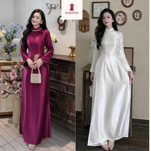 May include: A long-sleeved floor-length dress with a high collar, available in deep magenta or white satin fabric. The garment features a fitted bodice, a flowing skirt, and a small rectangular beaded handbag held in the front of the magenta version.