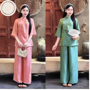 Könnte beinhalten: Zweiteilige Outfits in Rosa und Grün. Jedes Outfit besteht aus einem kurzärmeligen Oberteil mit Stehkragen und einer Hose mit weitem Bein. Das rosa Outfit wird mit einer kleinen gewebten Handtasche kombiniert. Das grüne Outfit wird mit einem Fächer kombiniert. Beide Outfits haben ein strukturiertes Aussehen.