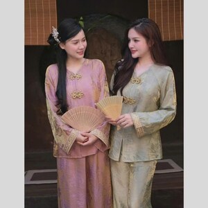 May include: Two women wearing traditional Asian-style outfits. One outfit is a light pink, the other is a light green. Both outfits have gold accents and are paired with matching fans. The outfits have long sleeves and are buttoned at the neckline.