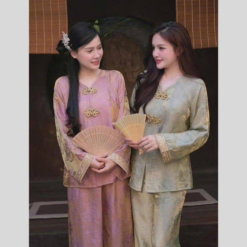 May include: Two women wearing traditional Asian-style outfits. One outfit is a light pink, the other is a light green. Both outfits have gold accents and are paired with matching fans. The outfits have long sleeves and are buttoned at the neckline.