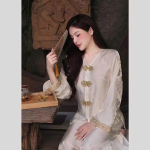 May include: Cream-colored silk pajama set with gold-colored floral embroidery and decorative clasps. The long-sleeved top has a V-neck and the pants are visible. A hand fan is held in the hand.