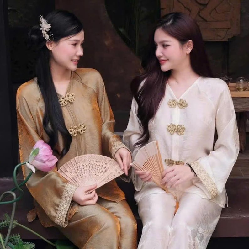 May include: Two women wearing traditional gold and cream-colored silk outfits, each holding a beige folding fan. The outfits feature ornate gold detailing on the sleeves and bodice. One woman has black hair, and the other has dark brown hair.