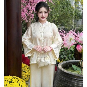 Op de afbeelding: Een cr&egrave;mekleurige tweedelige outfit met een top met mandarijnkraag en een broek met wijde pijpen. De top heeft delicate borduursels en kikkerknopen. Een kleine, bijpassende tas met een parelhandvat wordt voor de outfit gehouden. De achtergrond omvat roze bloemen en groen.