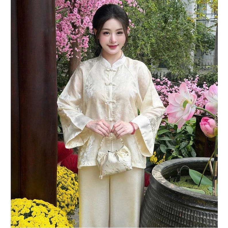 Op de afbeelding: Een cr&egrave;mekleurige tweedelige outfit met een top met mandarijnkraag en een broek met wijde pijpen. De top heeft delicate borduursels en kikkerknopen. Een kleine, bijpassende tas met een parelhandvat wordt voor de outfit gehouden. De achtergrond omvat roze bloemen en groen.