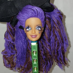 May include: A doll head with purple and brown hair, adorned with a large black bow. The doll has detailed eye makeup and a small heart design on its cheek. The head is attached to a green, textured stick.