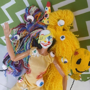 May include: A doll with a white face painted like a clown, wearing a yellow jumpsuit and an eye patch with a smiley face. The doll has multi-coloured yarn hair on one side and yellow yarn hair on the other, adorned with several plastic eyeballs.