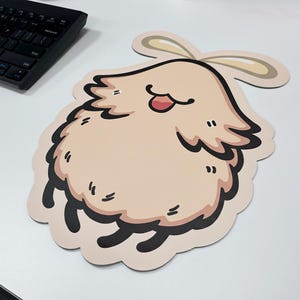 Pode incluir: Um mousepad bege com uma ilustração de desenho animado de uma criatura fofa e sorridente com uma língua rosa e detalhes em preto. O mousepad tem uma borda ondulada e duas formas bege semelhantes a orelhas na parte superior.