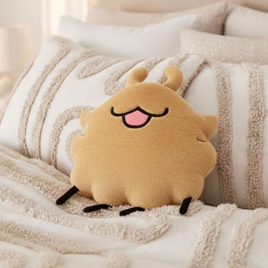 May include: A plush, tan-colored pillow shaped like a cartoonish creature with a smiling face. The pillow has small antennae and black accents for the eyes and feet. It rests on a bed with white and beige textured pillows and a matching comforter.