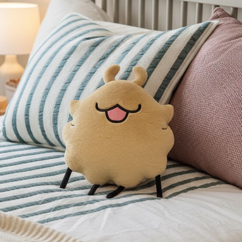 May include: A tan plush pillow shaped like a cartoon character with a smiling face and small black legs. The pillow has a pink tongue and is resting on a striped bedspread with pillows in the background.