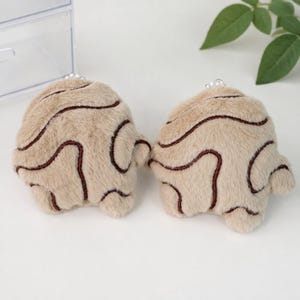 May include: Two plush, beige, fuzzy, rounded accessories featuring dark brown, wavy, embroidered lines across the surface. Each piece is topped with a small, white, spherical bead accent, creating a soft, textured appearance for these items.