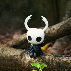 May include: A white and black figurine of a character with large, oval eyes and curved horns. The figure has a black cloak-like lower body and is posed on a tree branch. The background is blurred, showing a natural outdoor setting.