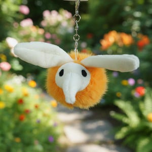 May include: A plush, orange bee-shaped keychain with white wings and a white face. The bee has black eyes and a long, white proboscis. The keychain is attached to a silver metal chain and clasp. The background is a blurred garden.