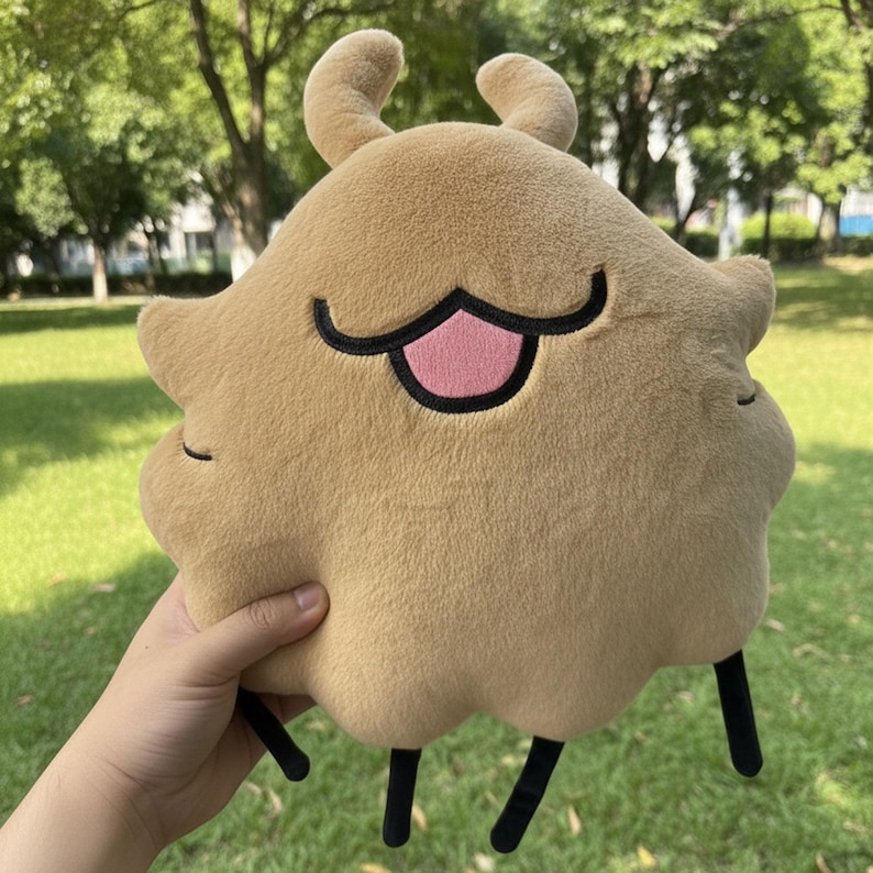 May include: A plush toy in the shape of a cartoon character, featuring a tan body with a black outline around the eyes and mouth, a pink tongue, and two small, curved antennae. The toy has black legs and is held in a hand.