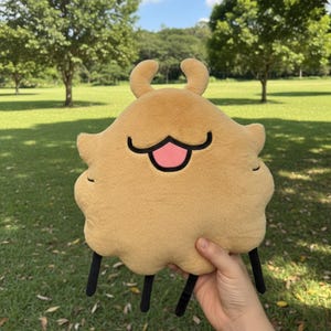 May include: A plush toy in the shape of a cartoon character with a tan body, black legs, and a smiling face with a pink tongue. The toy has two small, curved antennae and is held against a green grassy background.