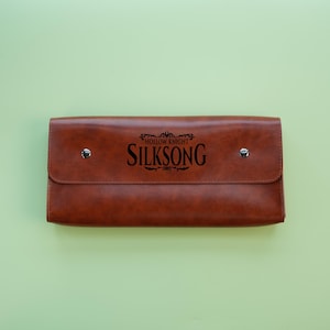 以下が含まれることがあります： 茶色の革製クラッチバッグ。フラップ開閉で、2つのシルバーのスナップ留めが付いています。「HOLLOW KNIGHT SILKSONG」の文字が黒でプリントされています。長方形のバッグは革製です。