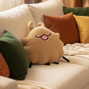 May include: A tan plush pillow shaped like a cartoon character with a smiling face, two small horns, and black antennae. The pillow is on a white sofa, surrounded by other colorful throw pillows in shades of green, orange, and yellow.