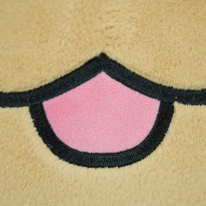 May include: Close-up of a plush toy's mouth, featuring a pink center and black outline against a tan background. The plush material has a soft, textured appearance, suggesting a cuddly toy.