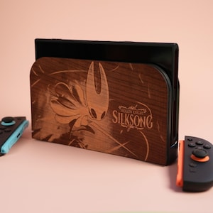 Puede incluir: Una base de Switch de madera con un diseño grabado con láser de Hollow Knight: Silksong. La base marrón presenta una imagen detallada del personaje. Se ven dos mandos, uno turquesa y negro, el otro negro y naranja.