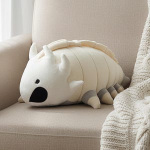 Könnte beinhalten: Ein cremefarbenes, insektenförmiges Kissen mit einem segmentierten Körper, kleinen Hörnern und grauen Beinen. Das Kissen liegt auf einem beigen Sessel neben einer cremefarbenen Strickdecke.
