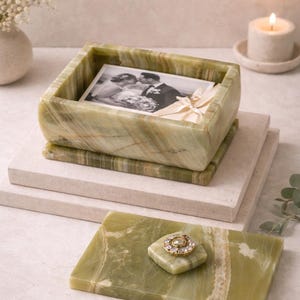 May include: A green onyx box with a photograph inside, resting on a stack of stone blocks. A matching onyx tray with a decorative gold and jewel accent is in the foreground. A lit candle and vase of flowers are in the background.