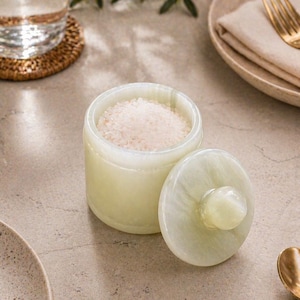 May include: A light green onyx salt cellar with a matching lid, partially open to reveal coarse salt. The cylindrical container sits on a neutral surface, suggesting a kitchen or dining setting. The lid is beside the container.