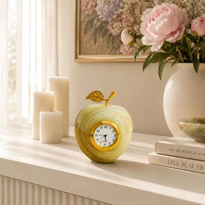 May include: An apple-shaped desk clock crafted from light green and white marbled stone, featuring a gold-toned metal stem and leaf accent. The clock face is circular with a gold rim, black numerals, and black hands, set against a smooth stone body.