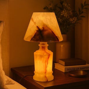 May include: A bedside lamp with a warm glow. The lamp features a rectangular lampshade and a base with a bottle-like shape. The lamp is made of a translucent material with brown and orange veining. The lamp sits on a dark wooden nightstand.