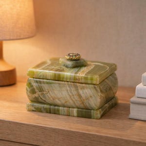 May include: A green onyx box with a gold-coloured knob on the lid. The box is rectangular with rounded edges and sits on a wooden surface. A lamp and books are in the background.
