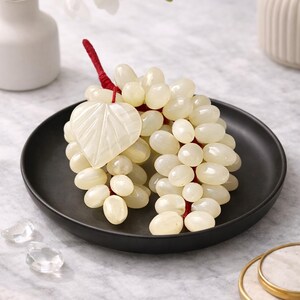 May include: A cluster of off-white, grape-shaped beads with a red stem and a leaf-shaped accent, presented on a black plate. The arrangement is part of a decorative display, with white orchids in a vase and other decorative elements.