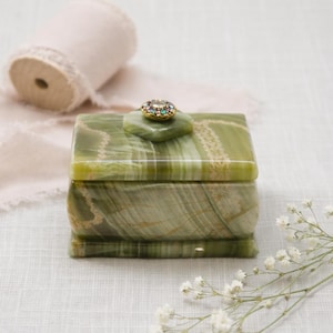 May include: A rectangular green onyx jewelry box with a decorative gemstone on the lid. The box has a marbled pattern and is set on a white surface. A roll of pink ribbon and white flowers are in the background.