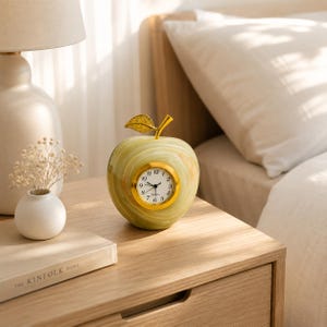 May include: A light green apple-shaped clock with a polished gold-tone stem and a circular clock face featuring black numerals and hands. The clock is crafted from a smooth, marbled stone material with subtle variations in color throughout the surface.