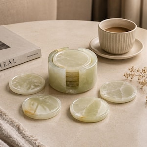 May include: A set of six green and white marble coasters with a matching holder. The coasters are round and have a natural, veined pattern. A cup of coffee sits on a saucer in the background.